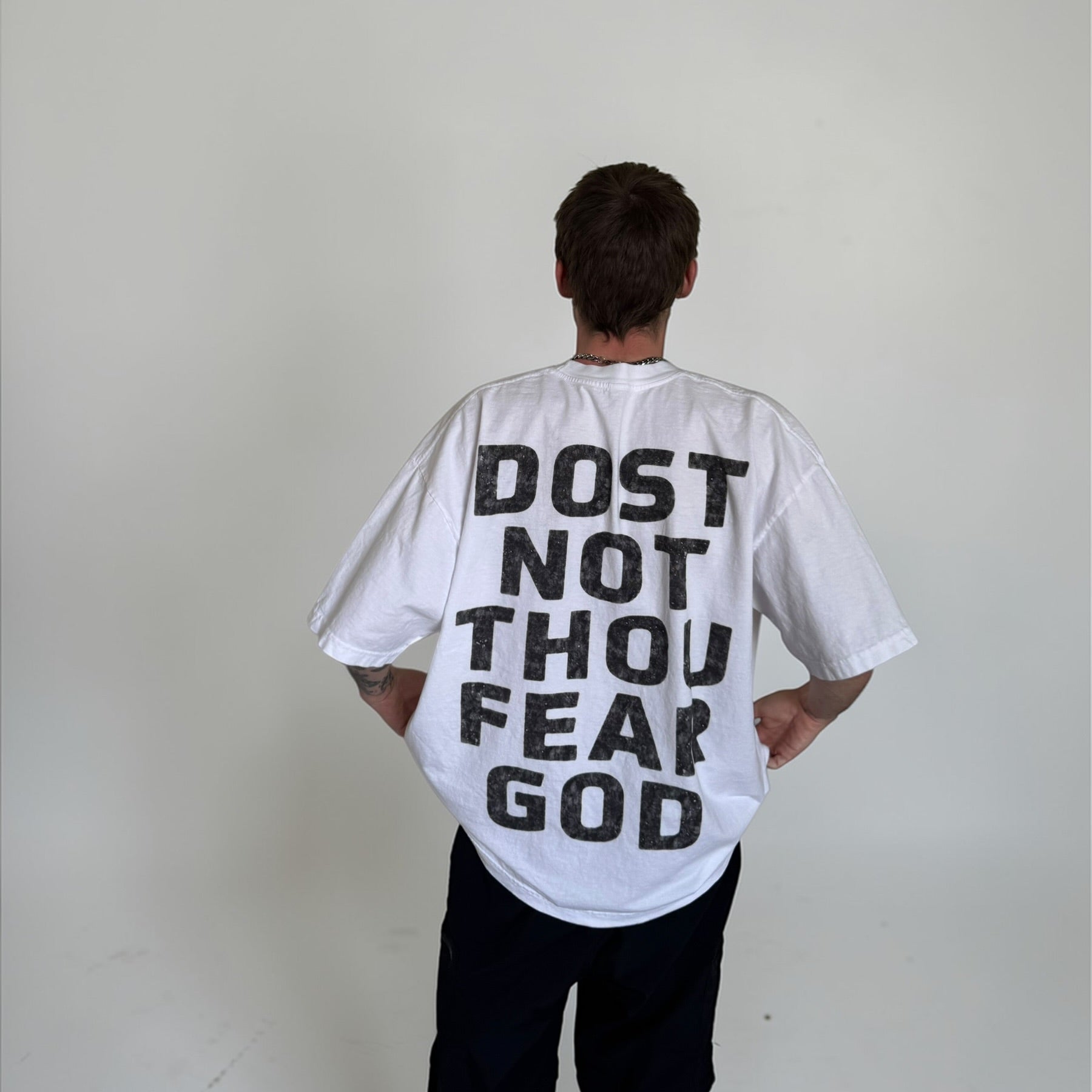 Person wearing a white t-shirt with 'DOST NOT THOU FEAR GOD' text on the back against a plain background